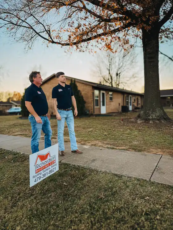 Two Richardson Roofing crew members in navy polos standing beside the company yard sign